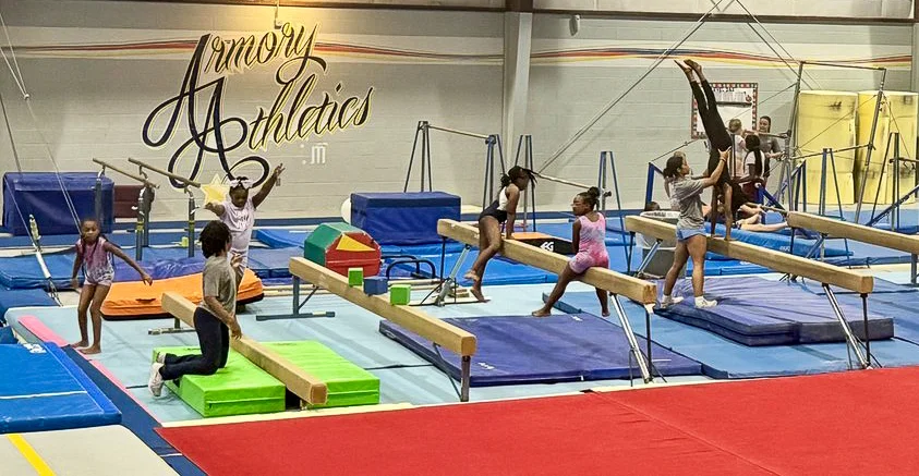 Recreational girls' class training on the balance beams beneath the Armory Athletics wall mural